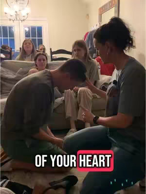 A group of people, indoors, with a man kneeling in prayer. A woman is touching the man's head and seems to be delivering a blessing while other people watch on a sofa. The hook text is in a pink box at the bottom of the image.