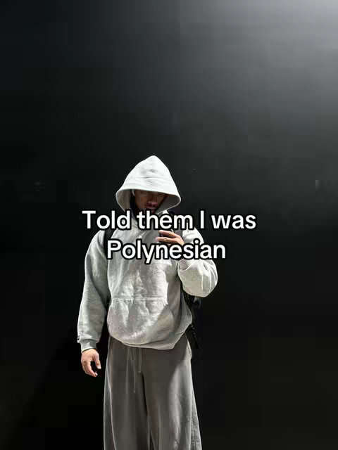 A person in a grey hoodie and matching sweatpants stands in front of a black backdrop. Text overlayed on the image reads 'Told them I was Polynesian'.