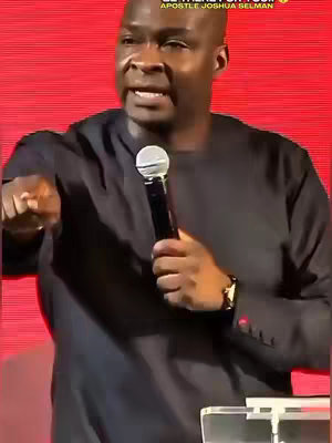 A man wearing a black long-sleeved shirt is standing in front of a red background. He's holding a microphone and pointing with his left hand. Text overlays are present at the top of the image, and a logo on the lower right corner.