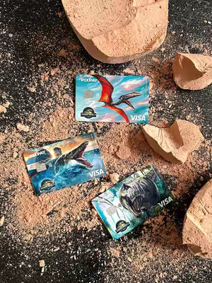 The image showcases three Pixpay Visa cards arranged on a dark, textured surface covered in fine brown particles, possibly sand or crumbled earth.  The cards feature dinosaur illustrations inspired by the Jurassic World franchise.  One card has a pterodactyl flying against a blue sky, another shows a creature in the water with its mouth open, and the third has a Tyrannosaurus Rex roaring.  The cards are interspersed with broken pieces of light-brown, rocky material, giving a prehistoric theme. The overall composition looks like a scene from a dig site or a museum display.