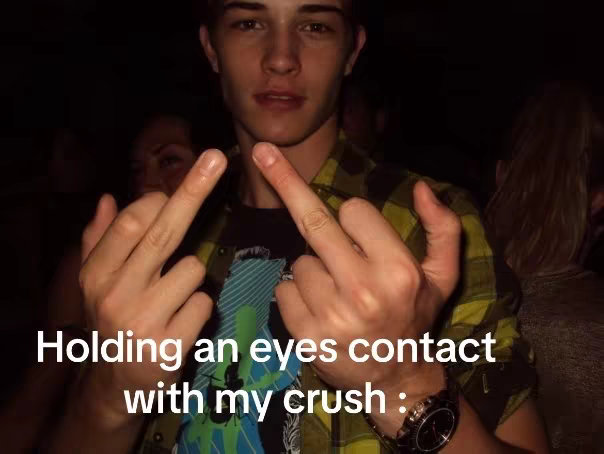 A young man with dark hair and a plaid shirt raises both hands, extending middle fingers on each hand. The background is blurred, suggesting a crowded, dimly lit environment. Text overlayed on the bottom describes the action.