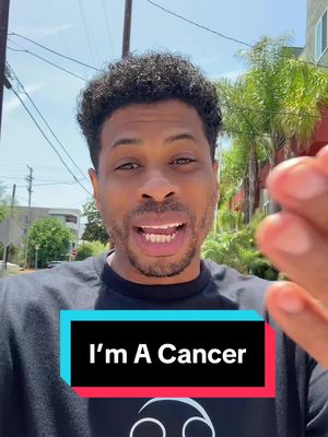 A selfie-style shot of a man in front of a residential street. He has short, dark, curly hair, a slight smile, and is looking directly at the camera. He is wearing a black t-shirt. The background contains power lines, buildings, and foliage. A colored text overlay at the bottom reads "I'm A Cancer".