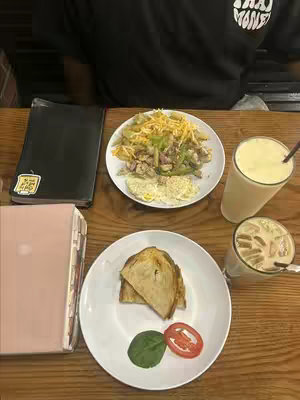 A photo showcasing a wooden table set for a meal, with two plates of food, two drinks, and two notebooks visible. One plate contains a breakfast with scrambled eggs, vegetables and what looks to be cheese, while the other has toast, a tomato, and a green leaf. There are two smoothies, one iced and one in a tall glass. A person wearing a black t-shirt with a graphic is visible.