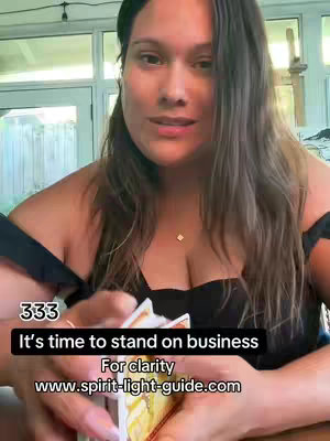 A woman is seen in a selfie-style shot, holding a deck of cards. The background has soft, green tones, suggesting an outdoor setting, with a window or door visible behind her. The woman has long, brown hair and is wearing a low-cut black top and a delicate necklace, with a focused expression on her face. Overlay text in a black rectangle provides guidance to stand on business.  The cards are in her hand, with some of the deck visible.