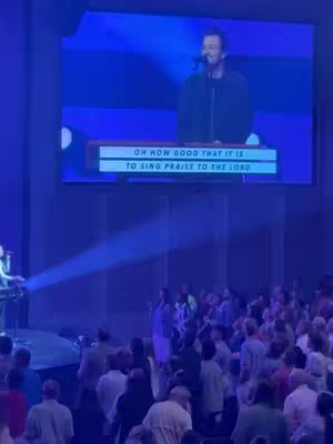 A live performance on stage with a man singing into a microphone. The backdrop is a large screen displaying lyrics, and the audience is visible in the foreground.