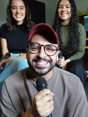 The cover image features three smiling individuals, with a man in the foreground holding a microphone, wearing a red cap and glasses. Two women are behind him, also smiling, holding microphones and sitting in the background. The overall composition is well-lit, with a focus on the people and their expressions.