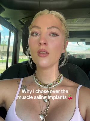 A selfie taken inside a car, featuring a fair-skinned woman with blonde hair and blue eyes. She is wearing multiple necklaces and a pink top. Text overlayed on the image reads 'Why I chose over the muscle saline Implants 🍒,' along with a cherry emoji.