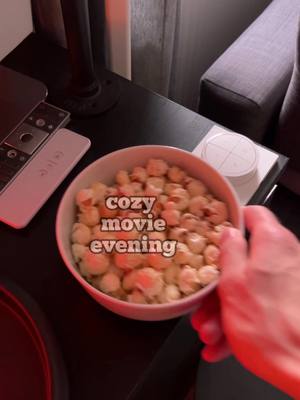 A low-angle shot shows a bowl of popcorn on a black surface, presumably a table. The popcorn fills the bowl, and the text overlay reads "cozy movie evening." A hand is reaching out to gently cradle the bowl. The details of the surroundings are slightly out of focus, suggesting the focus is on having a cozy movie night.