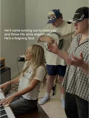 A group of three people, one woman and two men, are in a room. The woman is sitting at a piano, with long blonde hair, playing. One man in a baseball cap is standing, playing a banjo, and the other has an arm outstretched. Text overlayed in white says 'He'll come running out to meet you and throw His arms around you He's a forgiving God'.