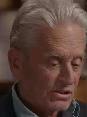 A close-up shot of an older man with gray hair, slightly furrowed brow, and a small smile. He appears to be looking down and to the side, possibly in thought or engaged in conversation. The lighting seems natural, and the man is wearing a collared shirt and a dark jacket or suit, suggesting a professional or interview setting.