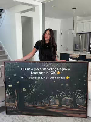 A woman stands in a well-lit, modern home, holding a framed piece of art that depicts a tree-lined road. The woman has long, dark hair and is wearing a black top. The background shows a staircase, a kitchen with white cabinets, and several windows.