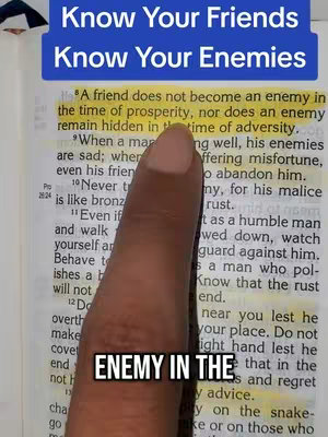 The image is a close-up of an open book, with a finger pointing at a highlighted passage and text overlay. The book has aged, off-white pages. The highlighted text is yellow, contrasting with the darker text surrounding it. A hand with visible skin tone with an out of focus background is placed in front of the highlighted text. There is also blue colored rectangular text box in the upper part with the caption Know Your Friends Know Your Enemies. Overlaid at the bottom of the image we can see the text ENEMY IN THE and some part of the letters. The text is in a bold, sans-serif font against a black rectangle.