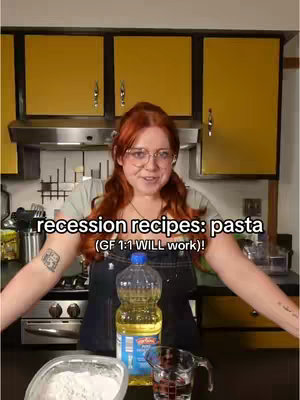 A woman with red hair and glasses stands in a kitchen, smiling at the camera. She is wearing a light green shirt and a black apron. Behind her are yellow cabinets. In front of her are bottles of oil, a bowl of flour, and a measuring cup of water.