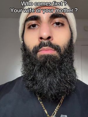 A close-up shot of a man with a thick, dark beard and a white beanie. He is wearing a black shirt and a gold chain. Text overlay at the top asks a question.