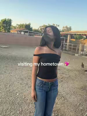 A woman stands in front of a rural backdrop wearing a black off-the-shoulder top and blue jeans. Her hair is blowing, partially obscuring her face. Text overlay reads 'visiting my hometown'. A pink heart emoji appears to the right of the text.