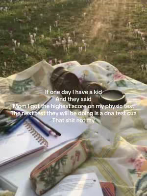 A photo of a picnic scene on a grassy area, with a patterned blanket spread out. On the blanket are several items including books, pens, a pencil case, and a coffee cup. The background consists of a blurred meadow dotted with white flowers, and the sunlight creates a warm atmosphere.