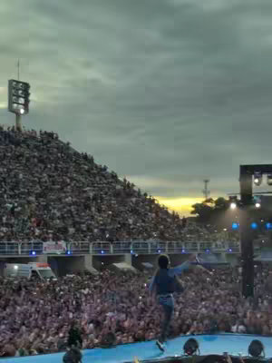 A live performance at a large outdoor event, likely a concert or religious gathering. A performer faces away from the camera towards a large crowd, with their arms outstretched. The crowd extends up a large, stadium-like structure. The sky is overcast, contributing to a sense of scale.