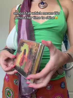 A person is shown from the waist up, holding a CD album in both hands. The person is wearing a green top and has a scarf around their neck. Text is overlaid on the image.