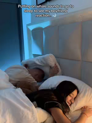 A woman is lying in bed, appearing to be asleep, while another person, possibly a man, is in bed beside her with their head covered by a pillow. The scene is dimly lit with a soft glow emanating from the headboard, creating a cozy, intimate setting.