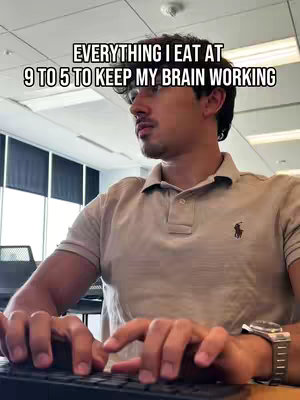 A man is shown in an office setting, sitting in front of a computer and typing on the keyboard. The main body of the image shows the man from the chest up. The man is wearing a light polo shirt with a polo symbol on the left side. His face is concentrated or focused. Above his head is a white text overlay that states "EVERYTHING I EAT AT 9 TO 5 TO KEEP MY BRAIN WORKING". The background is of a lit office with blinds.