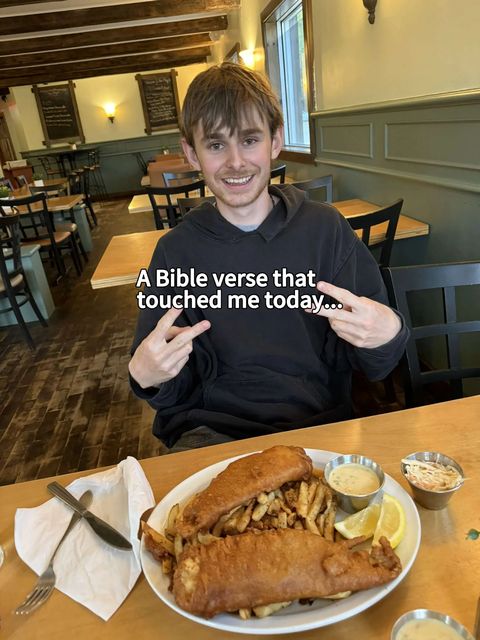 A young man with brown hair smiles at the camera, making a peace sign with his fingers while sitting at a restaurant table. In front of him is a plate of fish and chips, with a small container of sauce and a slice of lemon. The background has a neutral color scheme.