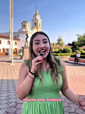 A woman stands in front of a building with a tall, ornate tower, holding a microphone in her mouth. The woman wears a green dress and smiles. The background includes a blue sky, trees, and a plaza with cobblestone.