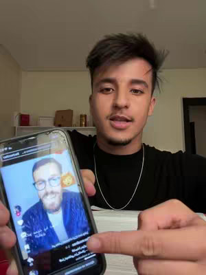 A young man in a black shirt holds up a smartphone displaying a video of another man, pointing at the phone with his right hand. The background is a lightly colored wall and some simple shelves.