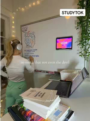 A woman, viewed from behind, stands with headphones on, writing on a whiteboard. The whiteboard has a diagram of a heart drawn on it. A laptop rests on the desk. The room is brightly lit and decorated with string lights.