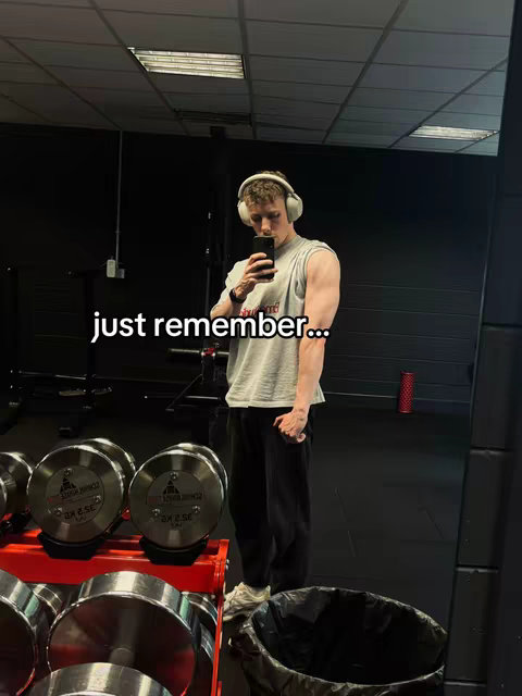 A man wearing a gray t-shirt, black pants, and white headphones is standing in a gym. He is taking a selfie with his phone, with the reflection of the gym equipment visible in a mirror in the background. The hook "just remember..." is overlaid on the image.