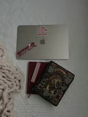 An overhead shot shows items resting on a white, textured surface, likely a bedspread or blanket. A silver laptop with an Apple logo and a sticker that says 'THE FUTURE IS BRIGHT' is positioned near the top. To the left of the laptop sits a pair of red-framed glasses. Below the laptop, there is a closed, maroon-colored journal next to a decorative, dark-covered book with ornate designs. The colors are muted and the lighting gives a soft feel to the image.