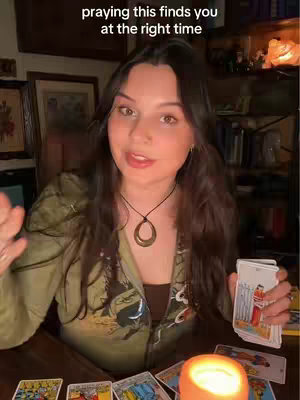 A woman with long dark hair is filming herself in a medium shot, holding a deck of tarot cards. There is a lit candle and other tarot cards on the table in front of her.