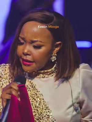 The image shows a woman with brown hair, wearing a gold-colored sequined top with a white collar, and holding a black microphone. The background is blurred, with some purple lighting visible.
