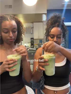 Two young women, likely on the younger side, are shown in the same room, possibly a cafe or eatery, each holding a clear plastic cup with a green beverage. The woman on the left looks directly at the camera with a slightly displeased expression, while the woman on the right stirs her drink. In the background, there are details of the interior, including a bright overhead light fixture, and a cabinet. The color scheme is warm with off-white hues. A text overlay is present at the bottom center of the image.