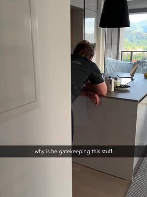 The image shows a person from the back, leaning against a countertop in a modern kitchen. The person is wearing a dark shirt and is partially obscured by a white wall. A laptop, coffee maker, and other items are on the counter. Natural light streams through a window in the background, partially obscuring the outside scenery.