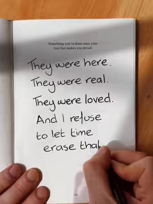 An open notebook page sits on a wooden surface. On the right side of the page, handwritten text is visible, seemingly written with a pen. The text is in a cursive style, and the words are arranged in a columnar format. The upper left of the page contains written text in a smaller size, while the bottom right corner showcases part of the page number.