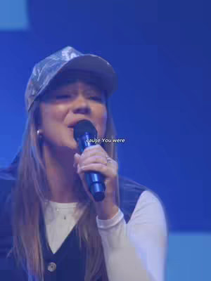 A woman with long blonde hair is singing into a microphone, wearing a camouflage baseball cap and a white long-sleeved shirt under a dark vest. The background is a blurred, blue-toned wash.