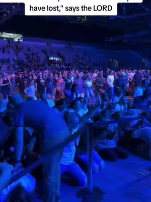 A large crowd of people in an arena-like setting. The image has a blue/purple color palette, likely due to stage lighting. The foreground is a bit blurred, with some people kneeling. In the background, many other people are standing and facing forward. At the top of the image, text is overlaid in a white box.