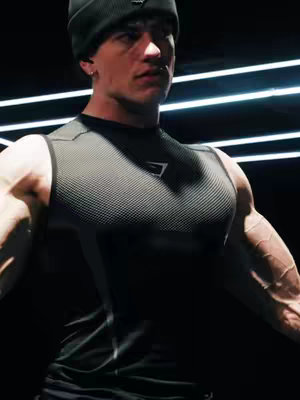 A muscular man wearing a black beanie and a sleeveless top is the focal point. His arms are flexed, showcasing defined muscles and visible veins. The backdrop is dark, with bright linear lights creating a geometric pattern behind him.