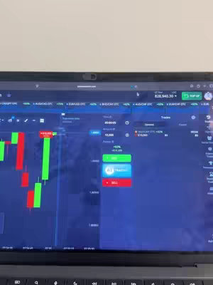 A close-up shot of a laptop screen displaying a trading platform interface. The screen shows a candlestick chart with red and green bars, indicating price fluctuations. There are various buttons and data points present, including "BUY" and "SELL" options, indicating a scenario involving binary options or similar financial trading. The laptop is on a desk, partially visible.
