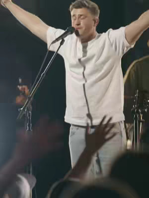Cover image of a man on stage with arms raised in worship. He wears a white shirt and light jeans, illuminated by stage lighting. The background is dark with other figures and musical instruments partially visible.