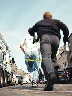 Two young men are running down a city street away from the camera, possibly in a moment of escape or excitement. The image is shot from a low angle, emphasizing the speed and movement of the runners with a slight fisheye effect. The street is lined with cars and buildings. The sky is slightly overcast.