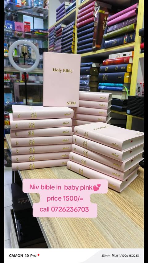 The image features a display of pink Bibles on a wooden table, likely in a bookstore setting. Several Bibles are neatly stacked, some upright and others lying flat, showing the covers. The background shows shelves filled with books. A text overlay in a pink rectangular shape provides pricing and contact information for this product, along with a heart emoji. The overall lighting is moderate, highlighting the pink color of the books against the wooden background.