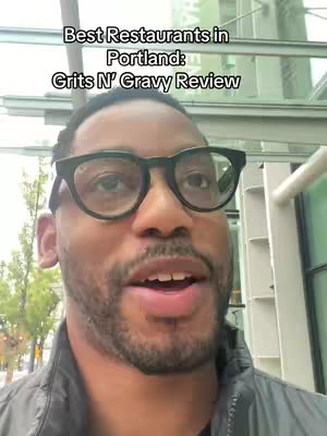 A dark-skinned man wearing glasses is the focus of a selfie. The background features a cityscape and glass windows. Text overlay at the top indicates the video topic: "Best Restaurants in Portland" and "Grits N' Gravy Review."