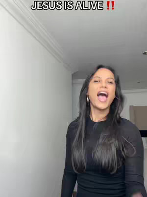 A woman with long, dark hair is centered in the frame, facing forward with an open mouth as if singing or speaking. Above her, text overlays read "JESUS IS ALIVE !!" in a bold, white font with red exclamation marks. The background is a simple, brightly lit interior with a smooth, plain white wall to the left and a suggestion of a doorway or hallway on the right. She is wearing a black top and large hoop earrings.