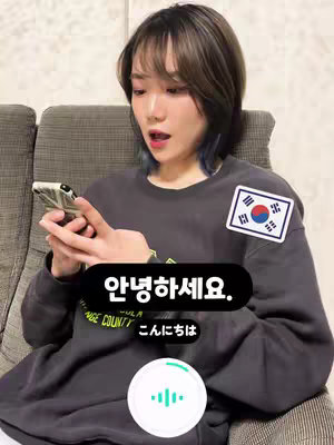 The cover image shows a young woman holding a phone, with an open-mouthed expression, amidst a dark gray sweatshirt. On her shirt is a South Korean flag sticker. The hook features Korean and Japanese text in a black box.