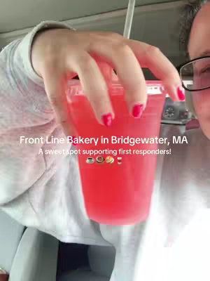 A selfie-style shot with the creator holding up a clear plastic cup filled with a bright red liquid. The creator's hand, with painted red nails, is visible. Text overlay says 'Front Line Bakery in Bridgewater, MA A sweet spot supporting first responders!'. The background is blurred, possibly inside a vehicle, with the creator's face partially visible, wearing glasses.