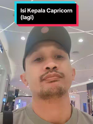 A selfie-style shot featuring a man in a black cap and a grey shirt at an indoor location, with an overlay of text. The man has a short beard and mustache. The background appears to be a shopping mall with bright lights. The overlay is a text box on top of the image, adding information to the image.