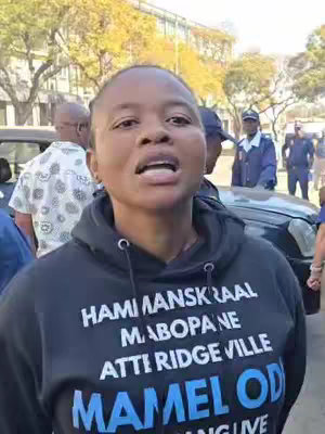 A woman with short hair wearing a black hoodie with white text is the main focus, set against an outdoor backdrop with other people and a vehicle. She is speaking, and the angle is a medium shot. The background includes a blurred street scene with additional individuals, buildings, and a car.