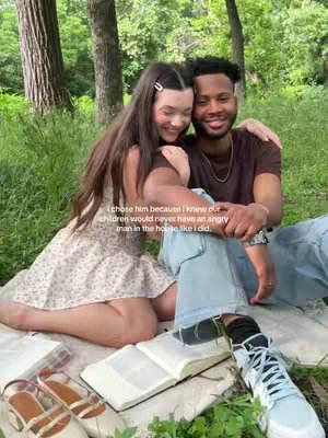 A young couple, a woman and a man, are sitting on a blanket in a grassy, wooded area. The woman is wearing a floral dress, and the man is in a brown shirt and jeans. They are smiling and embracing. An open book, shoes, and other items are on the blanket.