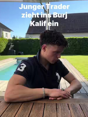 A young man sits outside at a wooden table with arms resting on the surface. A pool, green yard, and clear sky form the background. Text in a bubble-like font overlayed across the top reads 'Junge Trader zieht ins Burj', translating to 'Young Trader going into Burj'.